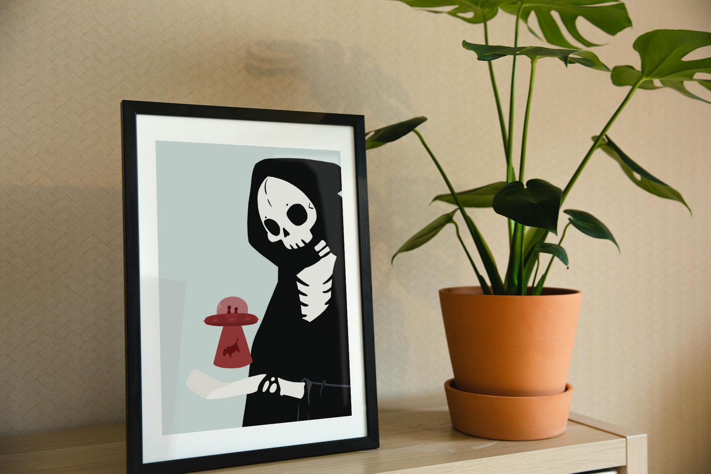 Framed art print on a shelf next to a house plant. The framed art print is of an illustration of a skeletal character with a red UFO abducting a cow hovering over its outreached hand in front of a light blue background.