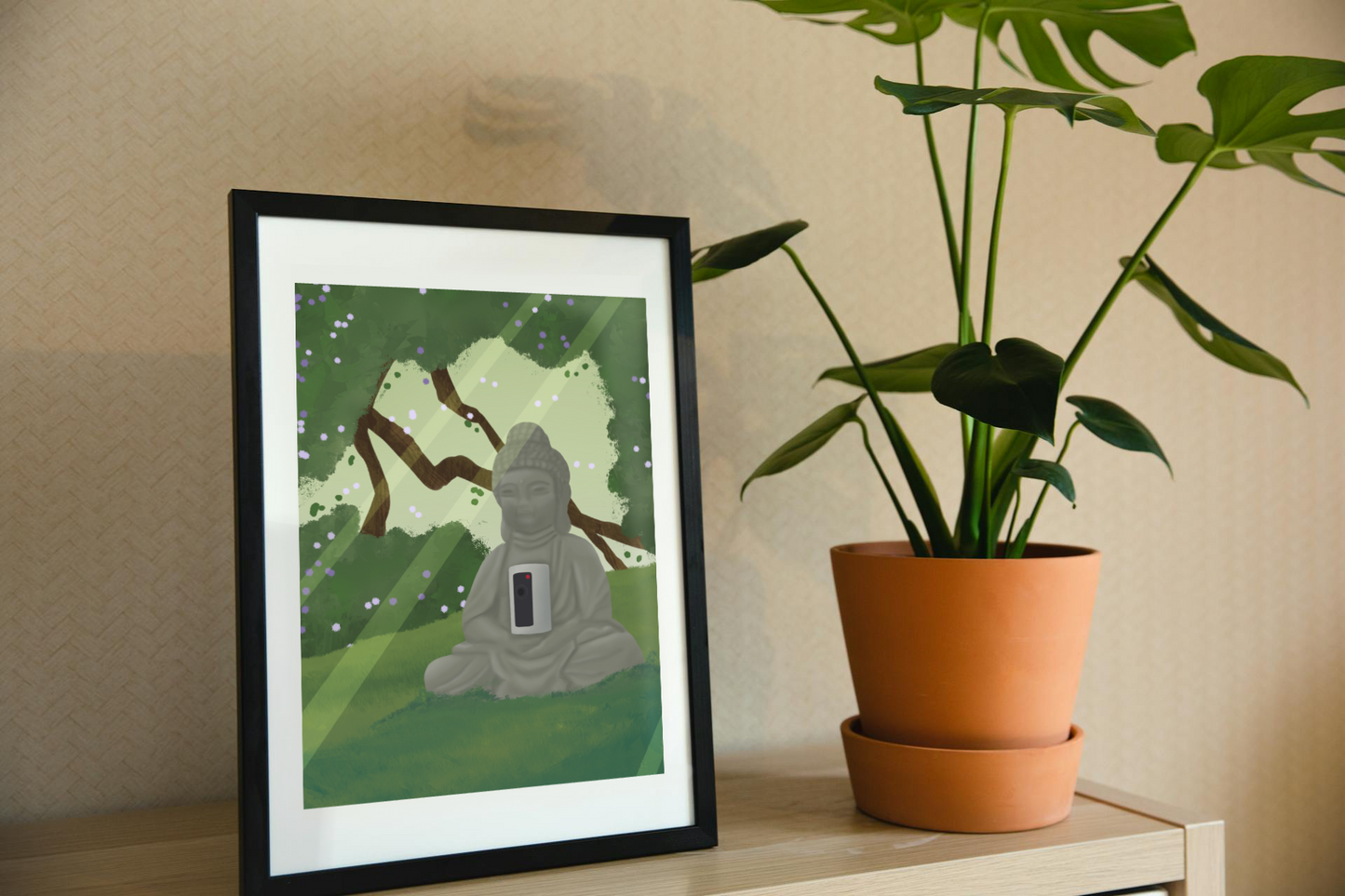 Framed art print on a shelf next to a house plant. The framed art print is of an illustration of a buddha statue with a security camera embedded into it, surrounded by trees, grass, and sunlight.