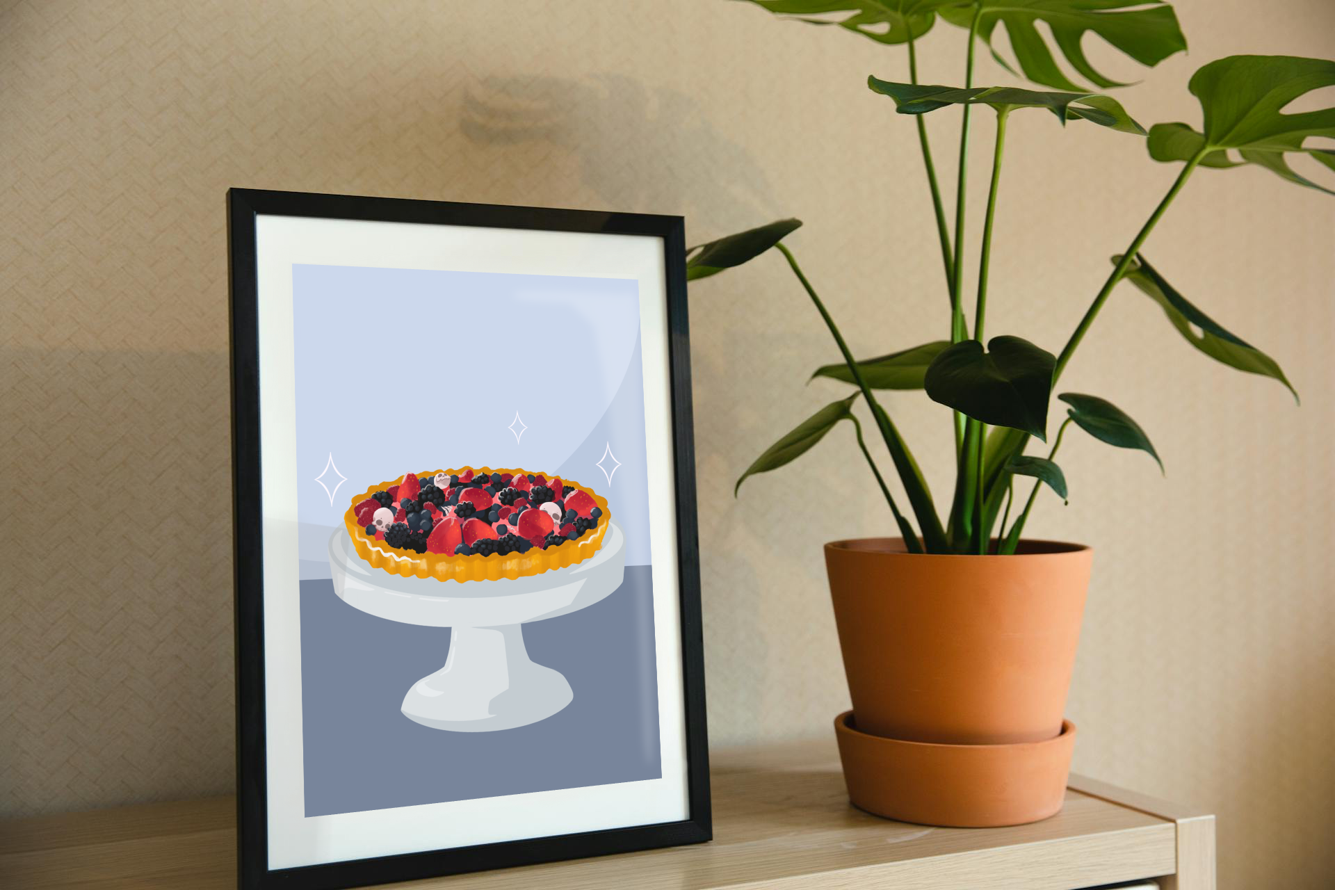 Framed art print on a shelf next to a house plant. The framed art print is of an illustration of a fruit tart filled with strawberries, blackberries, raspberries, blueberries, tiny skulls, and tiny hand bones on a gray cake stand against a light blue and dark blue background. Three sparkles surround the fruit tart.