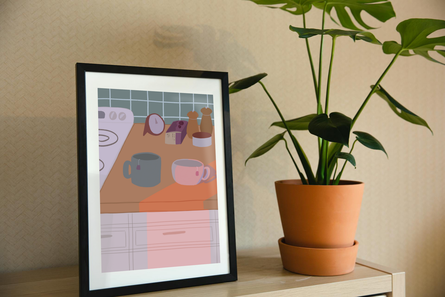 Framed art print on a shelf next to a house plant. The framed art print is of an  illustration of a kitchen counter with two mugs holding tea bags in cool evening lighting. In the background are two pepper grinders, a tea box, a salt dish, and a timer. On the left is a white stovetop, white cabinet drawers at the bottom, and green tiles for the backsplash.