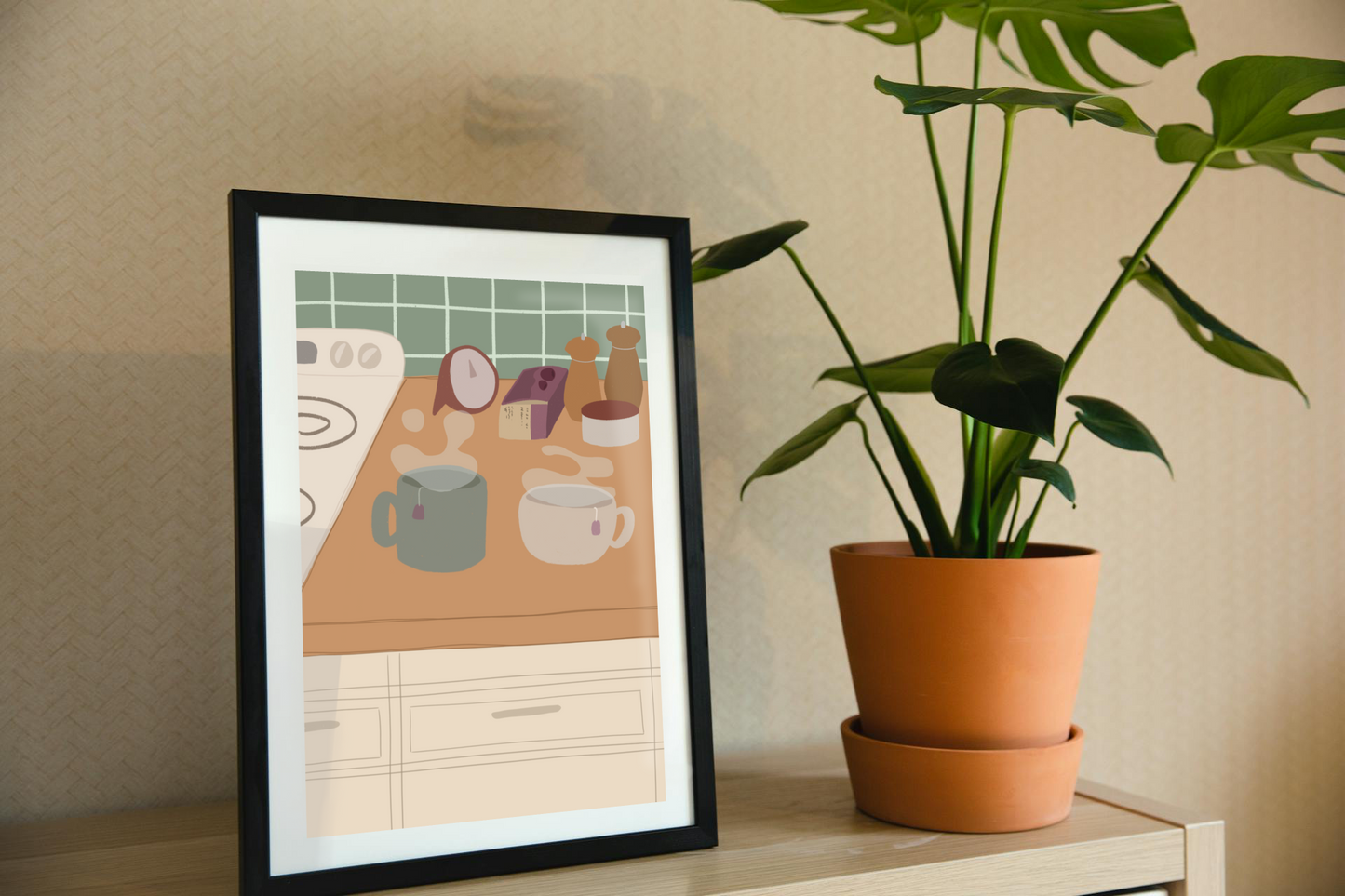 Framed art print on a shelf next to a house plant. The framed art print is of an illustration of a kitchen counter with two steaming mugs holding tea bags in morning lighting. In the background are two pepper grinders, a tea box, a salt dish, and a timer. On the left is a white stovetop, white cabinet drawers at the bottom, and green tiles for the backsplash.