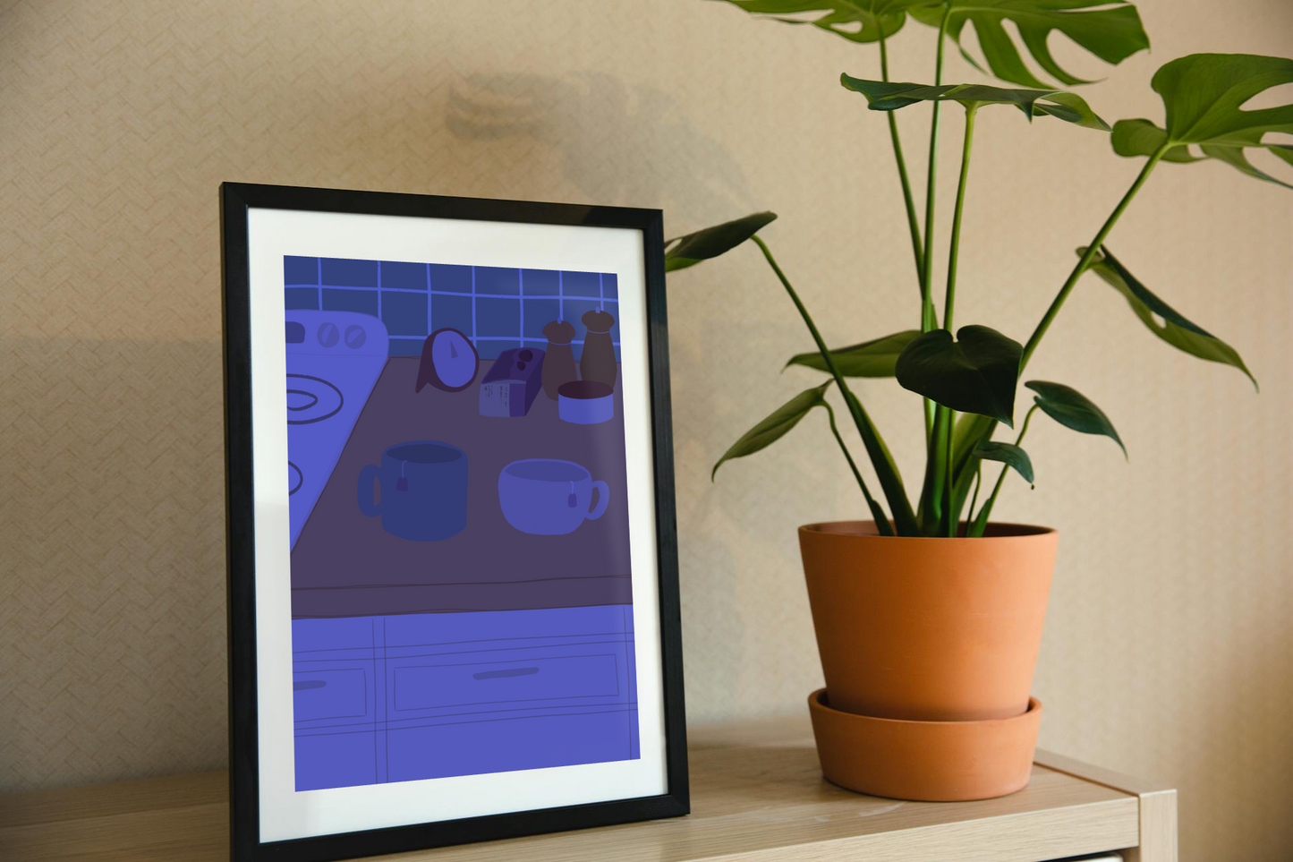 Framed art print on a shelf next to a house plant. The framed art print is of an illustration of a kitchen counter with two mugs holding tea bags in blue night lighting. In the background are two pepper grinders, a tea box, a salt dish, and a timer. On the left is a white stovetop, white cabinet drawers at the bottom, and green tiles for the backsplash.