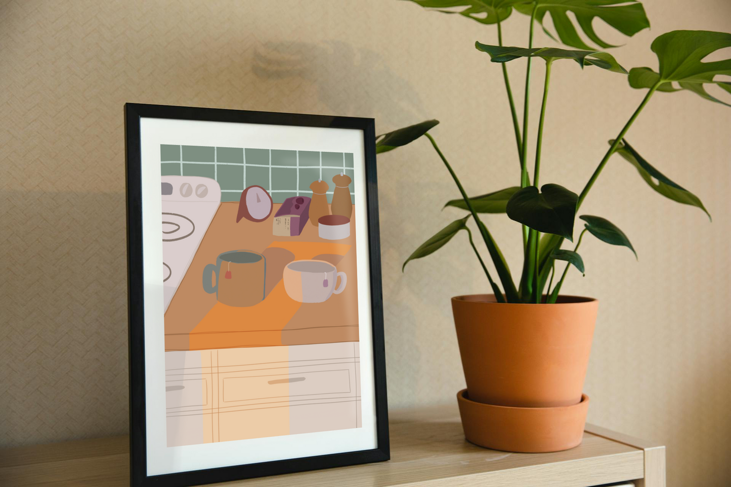 Framed art print on a shelf next to a house plant. The framed art print is of an illustration of a kitchen counter with two mugs holding tea bags in warm afternoon lighting. In the background are two pepper grinders, a tea box, a salt dish, and a timer. On the left is a white stovetop, white cabinet drawers at the bottom, and green tiles for the backsplash.