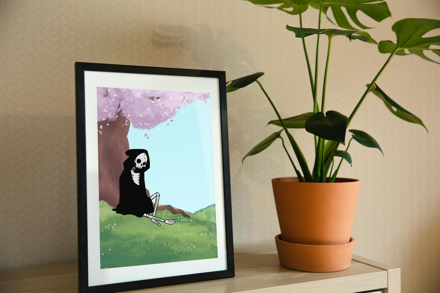 Framed art print on a shelf next to a house plant. The framed art print is of a skeleton under a cherry blossom tree with a light blue sky in a hilly field next to a potted plant on a shelf.