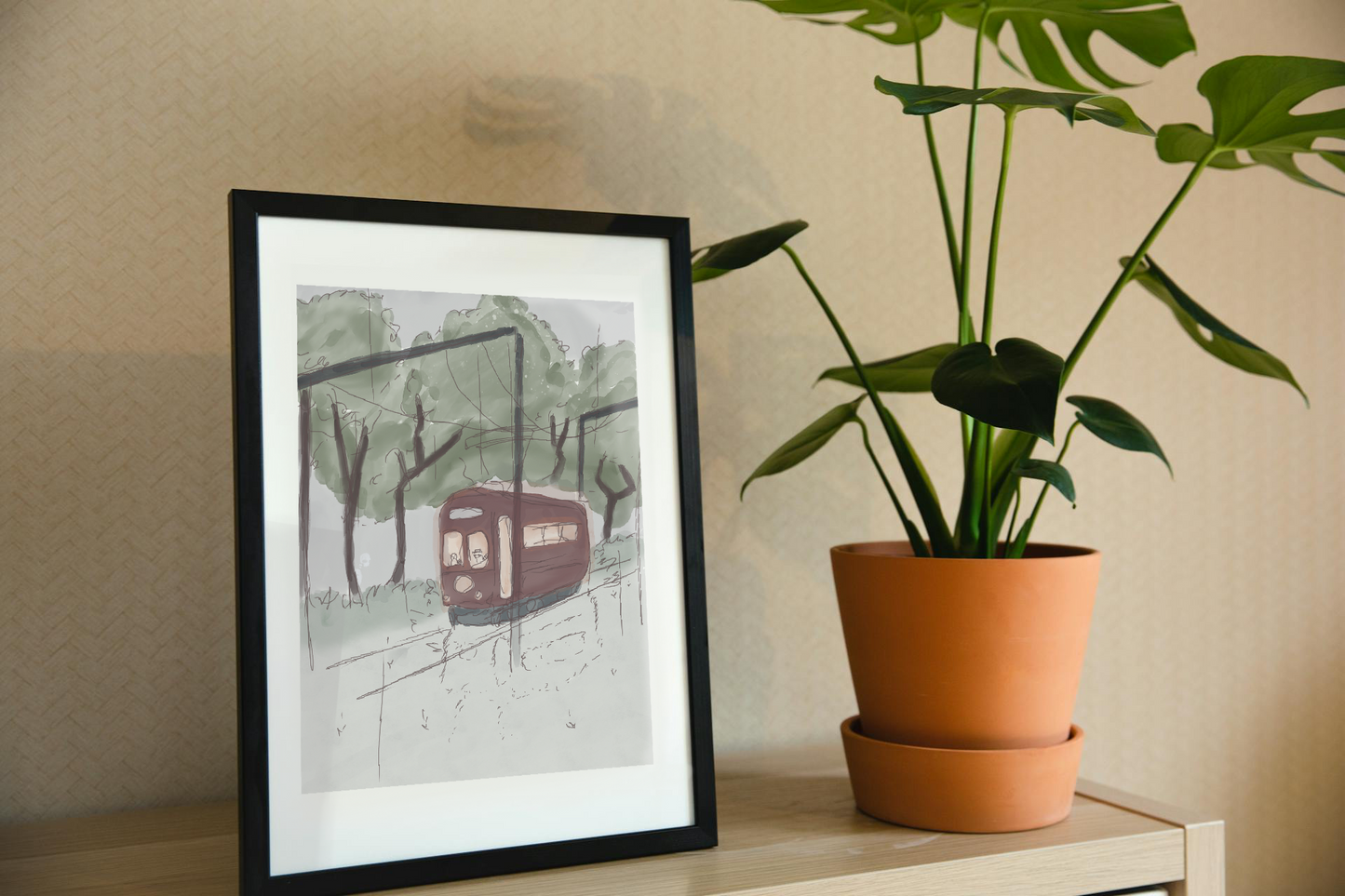Framed art print on a shelf next to a house plant. The framed art print is of an illustration of a watercolor-style tram in a forest with trees, a cloudy sky, and light rain. A beaver and raccoon drive the tram, while a skeleton hidden in the trees watches as it passes by.